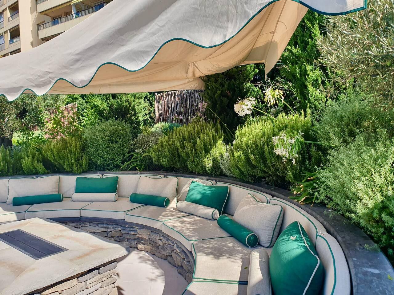 Salotto esterno circolare in pietra con cuscini verdi sotto vela ombreggiante beige, giardino mediterraneo